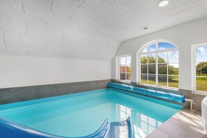 A swimming pool with a blue edge, a bench, and a view of the outdoors through large windows.