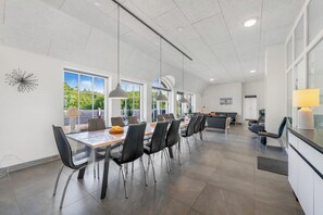 A spacious dining area with a long table, multiple chairs, pendant lighting, and large windows offering a view of greenery.