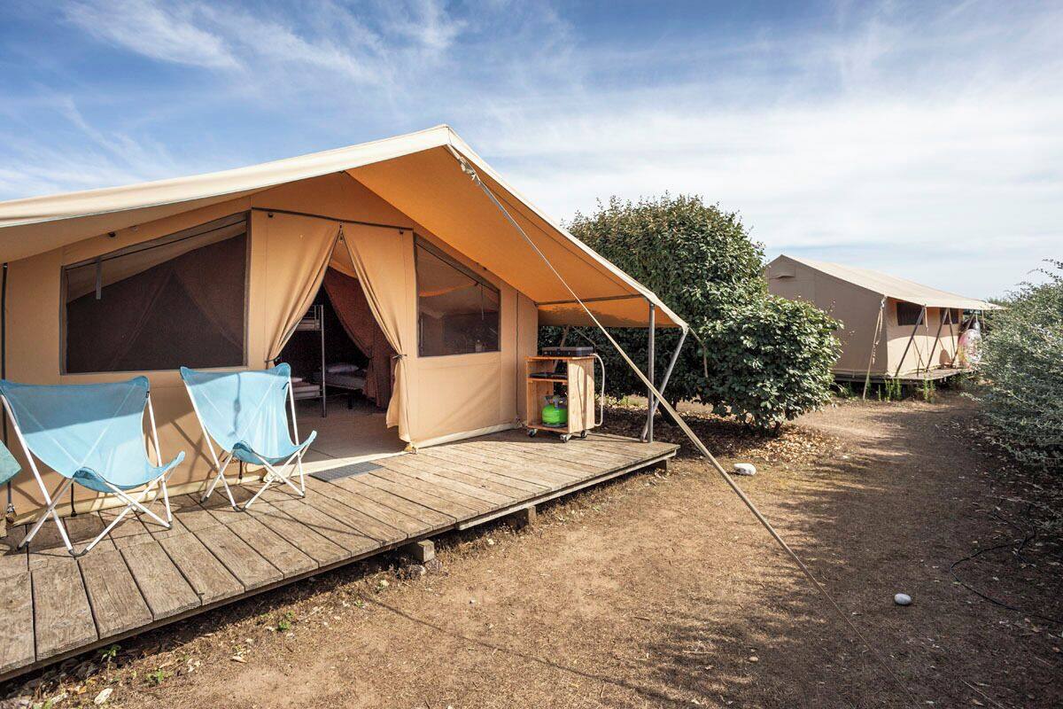 Un terrain de camping avec une tente, deux chaises pliantes bleues et une petite terrasse en bois.