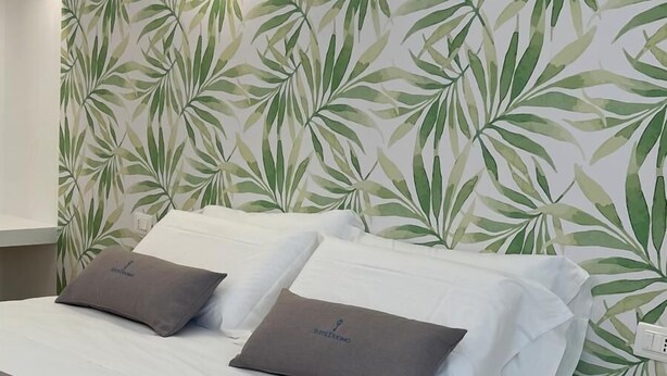 A neatly made bed with white linens and two pillows, a wall with a leafy pattern, and a round table with a geometric design. - Suite Duomo Tropea (Tropea)