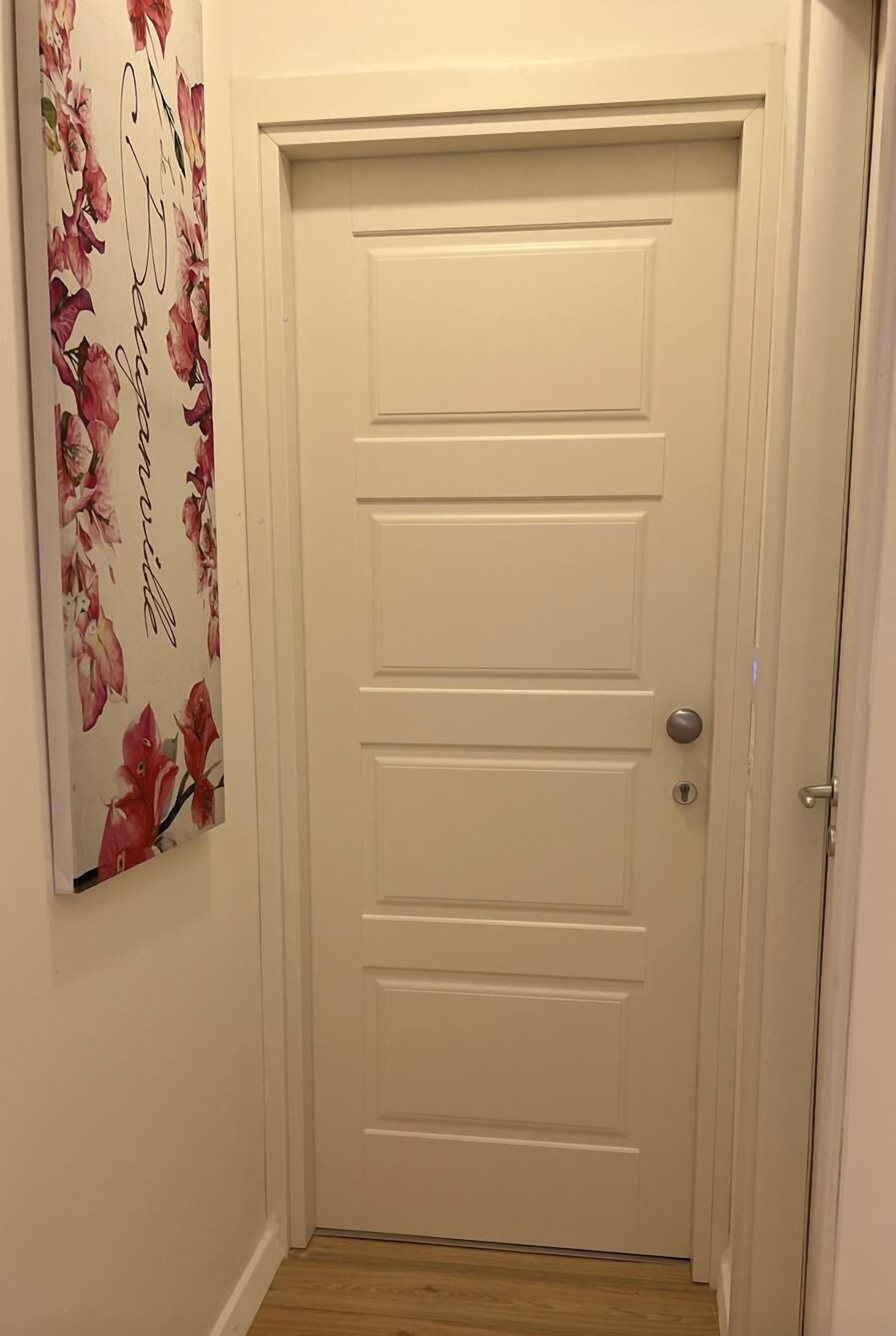 A white door with a keyhole and a doorknob, a floral wall decoration to the left, and a wooden floor.