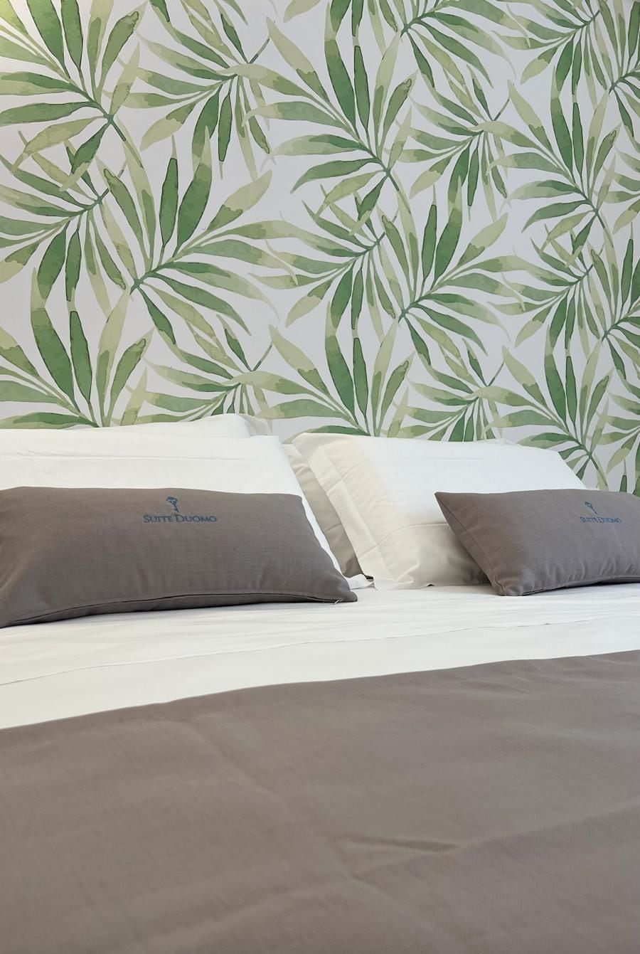Una cama con dos almohadas, cabecera blanca y pared con diseño de hojas.
