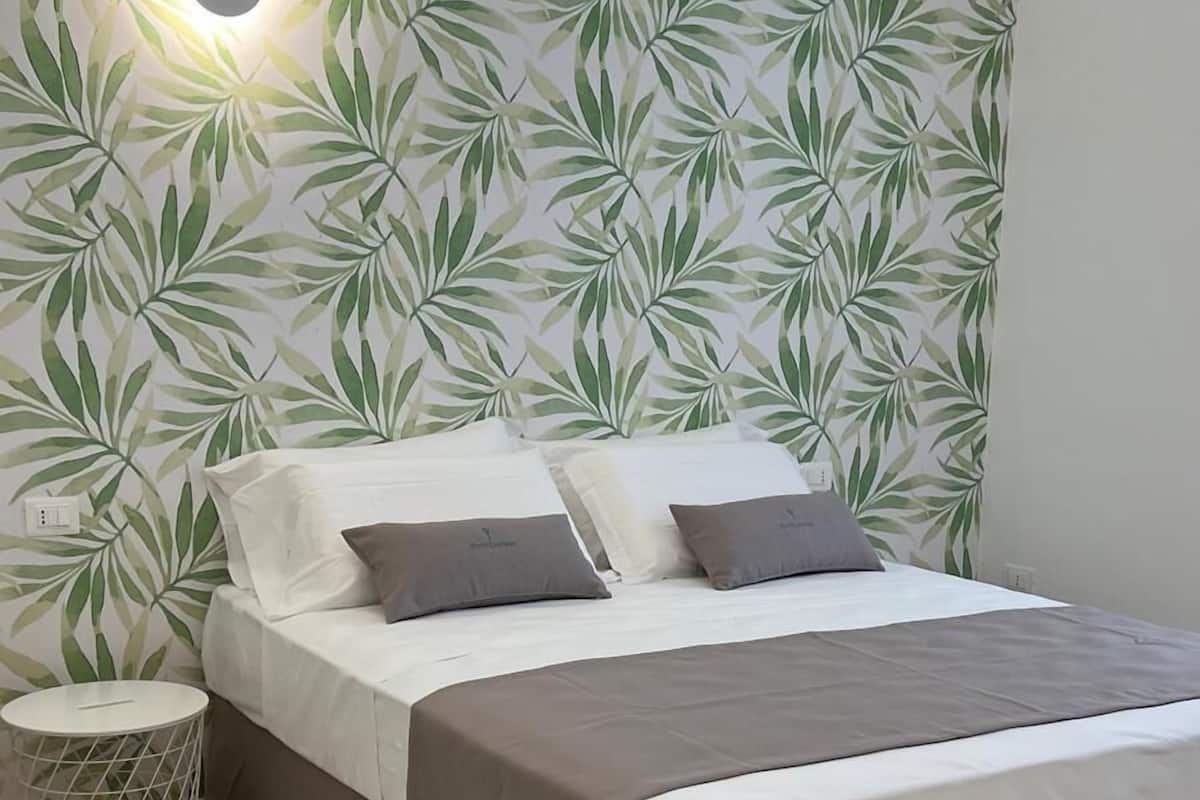 A neatly made bed with a white and brown bedspread, a small round nightstand with a white lamp, and a wall with a leafy pattern.