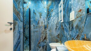 Un baño con cabina de ducha de vidrio, inodoro blanco y un lavamanos ovalado grande sobre encimera de mármol.
