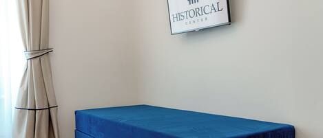 Ein Zimmer mit einem Bett, das blaues Bettzeug trägt, einem an der Wand befestigten Schild mit der Aufschrift "HISTORICAL CENTER" und einem am unteren Rand gebundenen Vorhang.