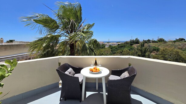 Un balcon agrémenté de fauteuils en osier et d’une petite table, offrant une vue sur un paysage côtier avec des palmiers et des maisons.