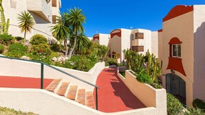 Un chemin mène à un bâtiment dont le sol est recouvert de tuiles rouges et les murs sont blancs, et qui est entouré de cactus et de palmiers.