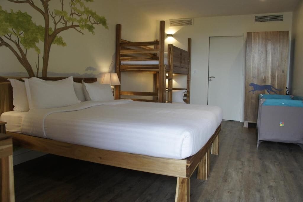A bedroom with a wooden bed, a nightstand, a desk, a wall-mounted shelf, and a door with a wooden panel.