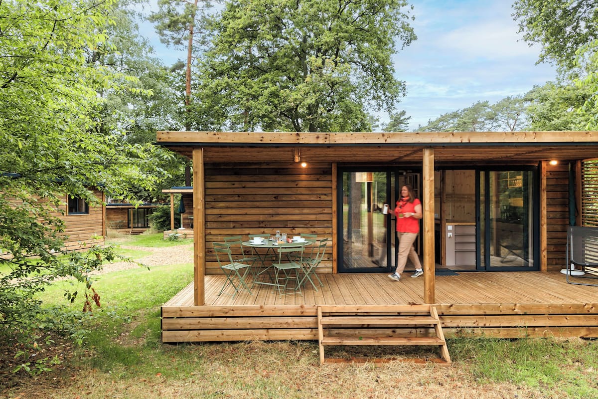 A wooden cabin with a deck, a dining table with chairs, and a glass door leading to an interior space.