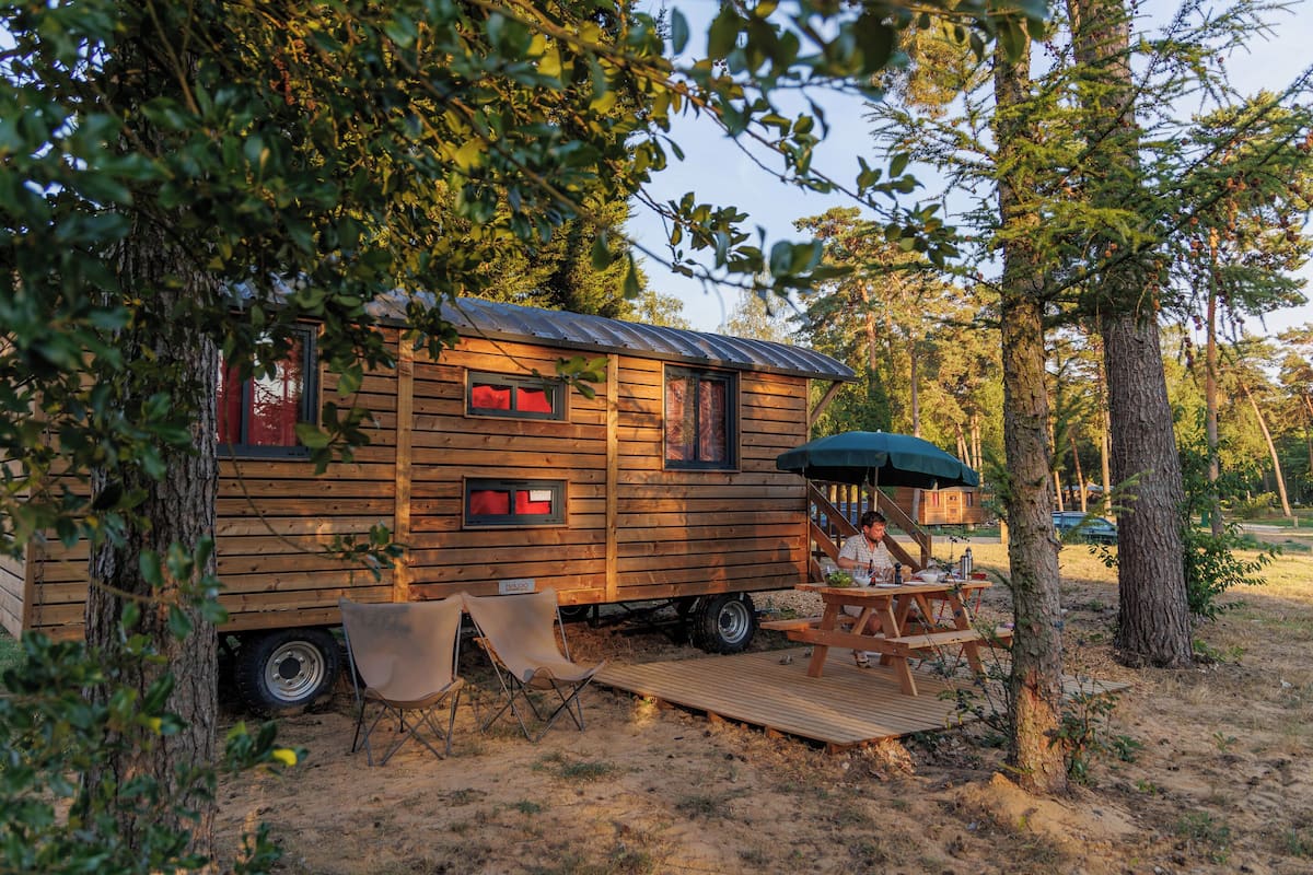 A wooden cabin with a picnic table and chairs, surrounded by trees.