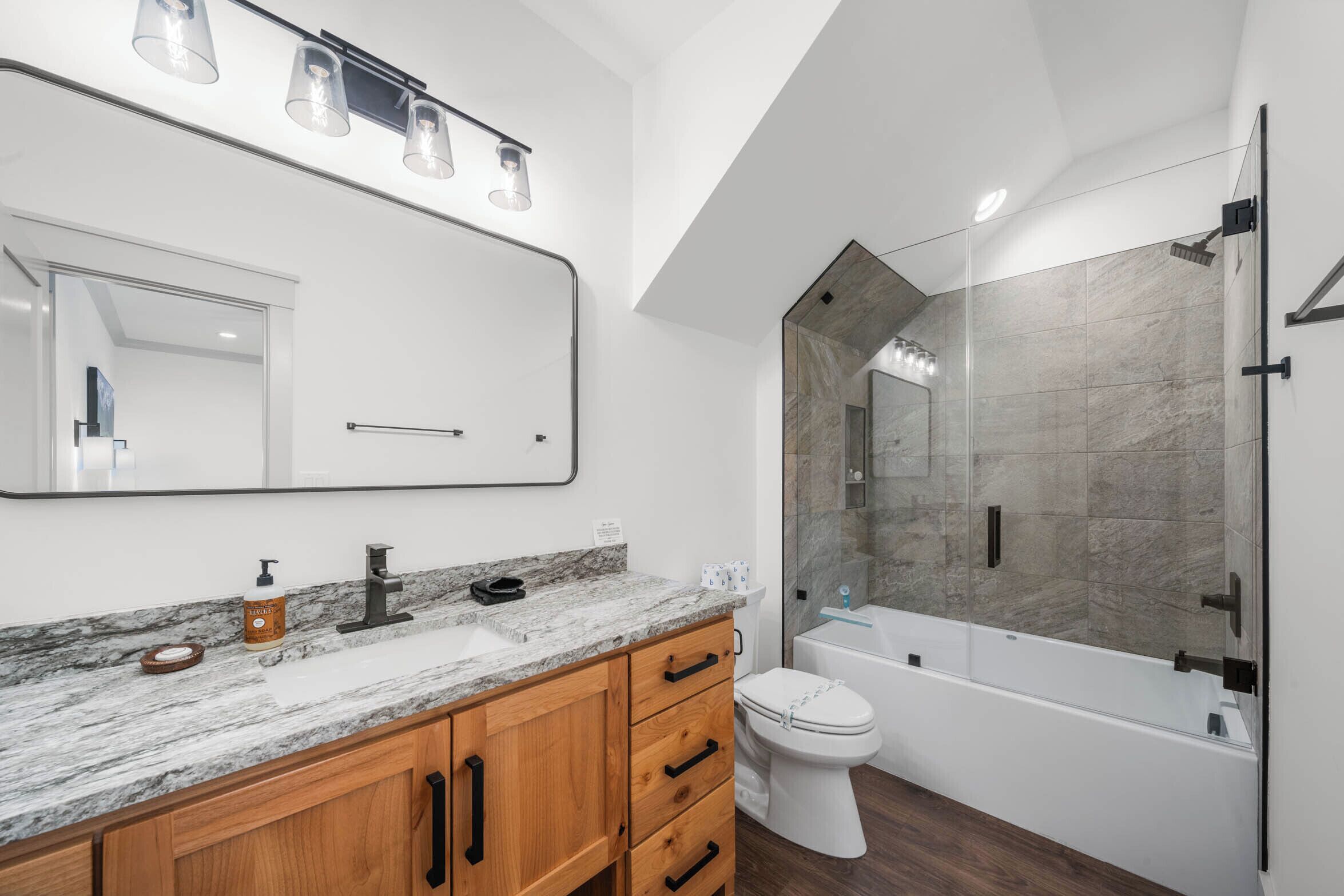 Une salle de bain moderne avec un comptoir en marbre, des meubles en bois, un grand miroir, des toilettes et une douche séparée par une paroi de verre.