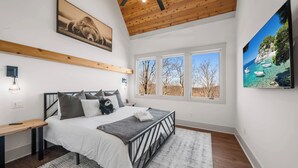 A bedroom with a bed, a bedside table, a wall-mounted TV, and a window with a view of trees and a clear sky. - Modern Mountain Lodge with Great Amenities (Boone)