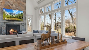 A living room with a stone fireplace, a large TV mounted above it, and a wooden dining table with a decorative centerpiece. - Modern Mountain Lodge with Great Amenities (Boone)