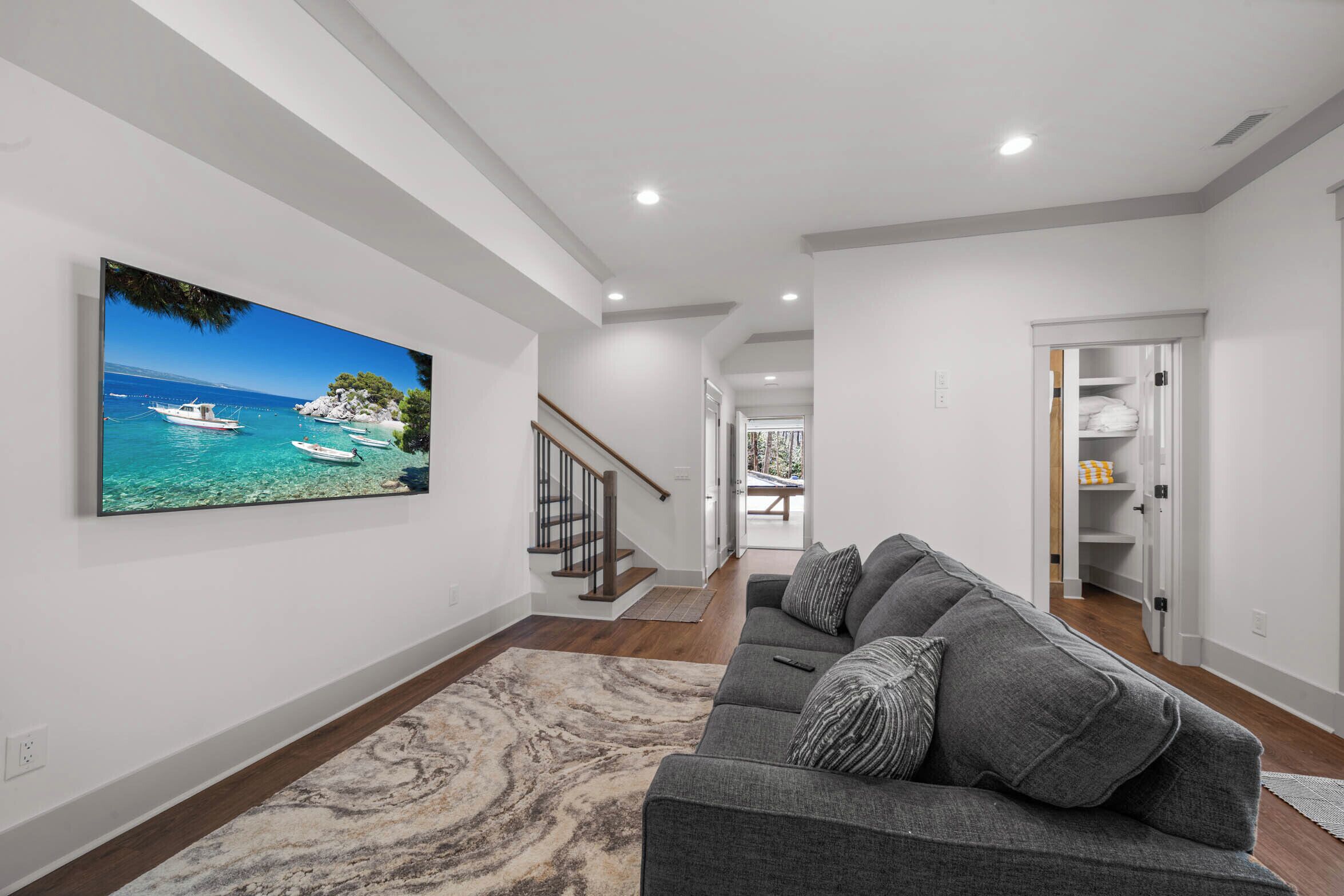 A modern living room with a large flat-screen TV, a gray sofa, and a wooden staircase.