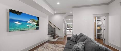 A modern living room with a large flat-screen TV, a gray sofa, and a wooden staircase.