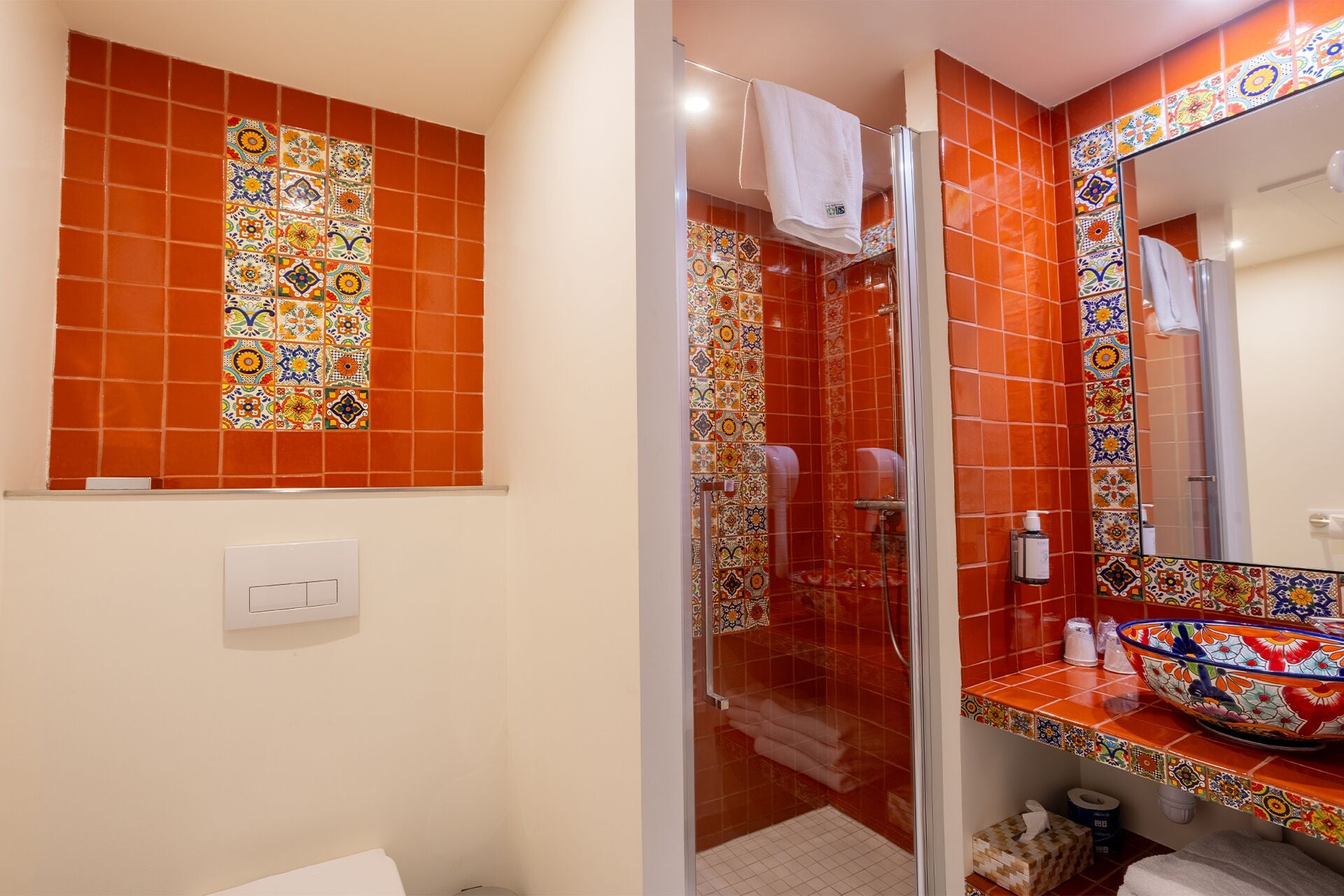 A bathroom with a glass shower enclosure, a tiled wall with a colorful pattern, a sink with a decorative bowl, and a mirror.