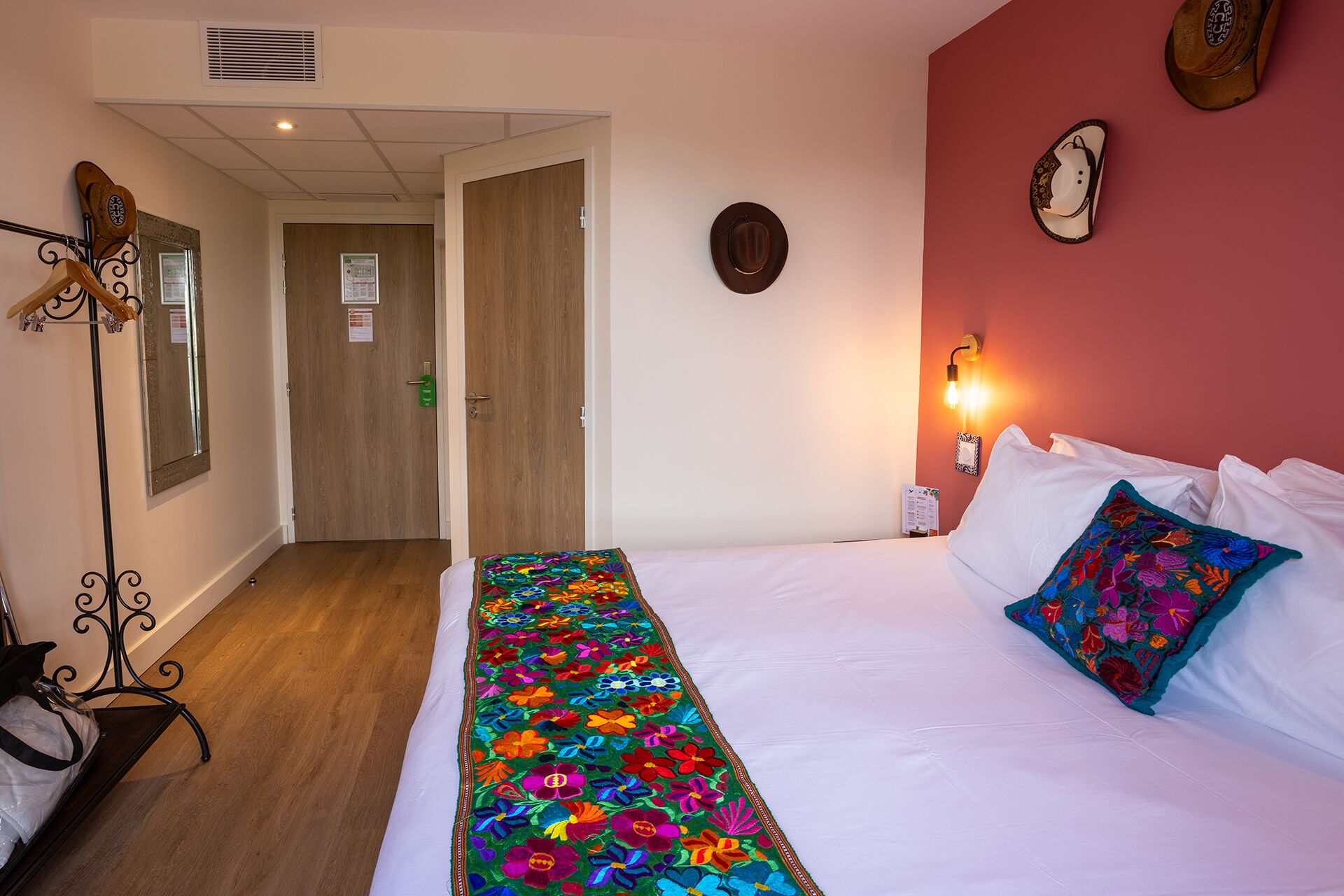 A hotel room with a bed, a colorful embroidered blanket, a wooden door, a coat rack, and a wall clock.