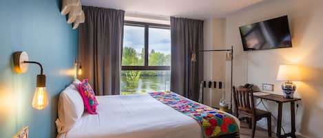 Ett hotellrum med en säng, ett skrivbord, en stol, en tv och ett fönster med utsikt över grönområden.