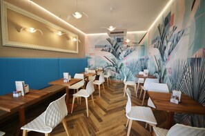 A restaurant interior with wooden tables and white cushioned chairs, a blue cushioned bench, and a mural of tropical plants on the wall. - Hotel Olimpia - Family Suite Standard (Bibione)
