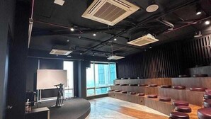 Uma sala de conferências moderna com palco curvo, fileiras de assentos e uma grande janela que proporciona luz natural.