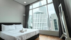 Quarto de hotel moderno com cama grande, ventilador de teto e vista para prédios da cidade através de janelas amplas.