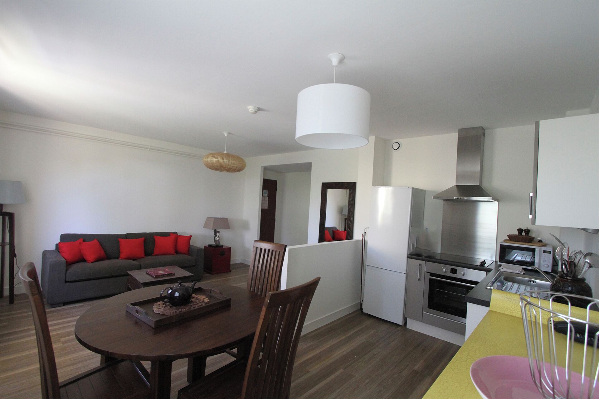 A modern kitchen with a stainless steel range hood, a built-in oven, and a microwave. There is a dining area with a wooden table and chairs, and a sofa with red cushions.