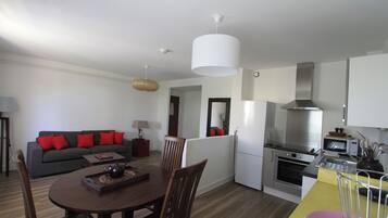 A modern kitchen with a stainless steel range hood, a built-in oven, and a microwave. There is a dining area with a wooden table and chairs, and a sofa with red cushions.
