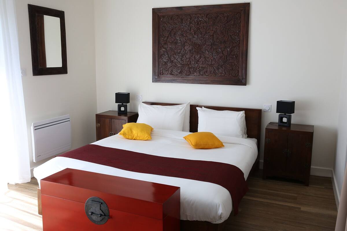 A hotel room with a bed, two bedside tables with lamps, a red cabinet, a large mirror, and a decorative wall panel.