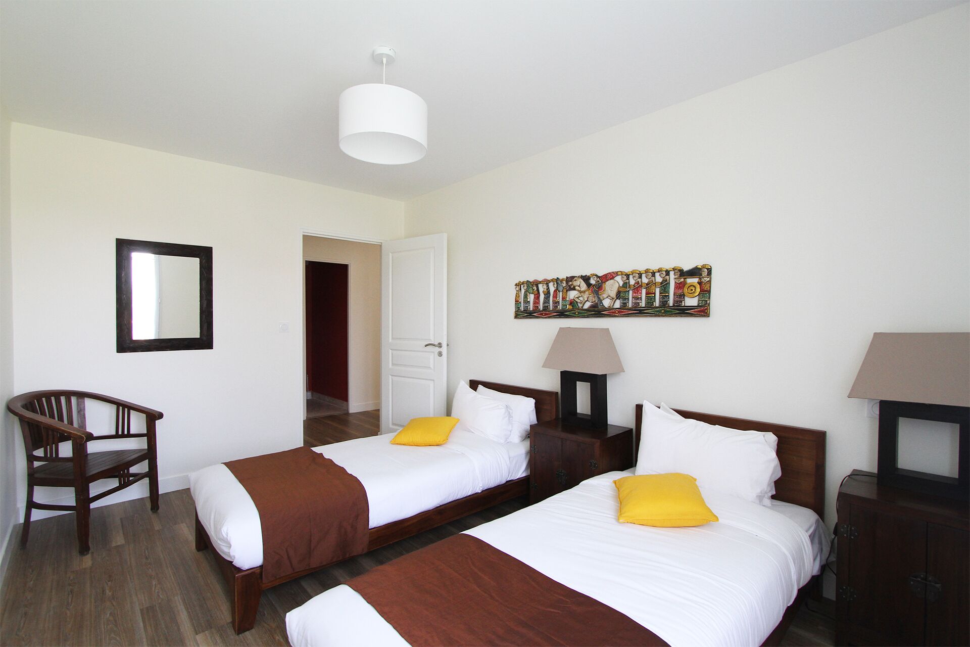 Quarto de hotel com duas camas, uma cadeira, uma porta, um espelho, uma obra de arte na parede e um abajur.