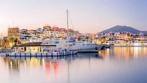 Un port de plaisance où est amarré un grand yacht, entouré de bâtiments dotés de balcons, avec une montagne en arrière-plan.