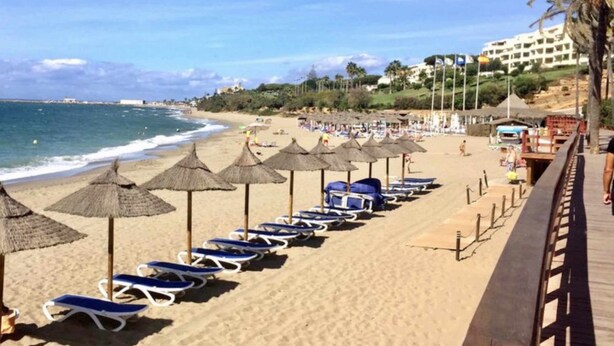 A beach with rows of sun loungers and thatched umbrellas, a wooden boardwalk, and a building in the background.