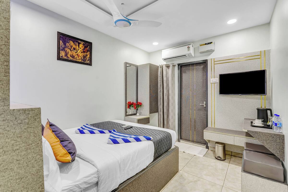 A hotel room with a bed, a desk, a TV, an air conditioner, a mirror, and a ceiling fan.