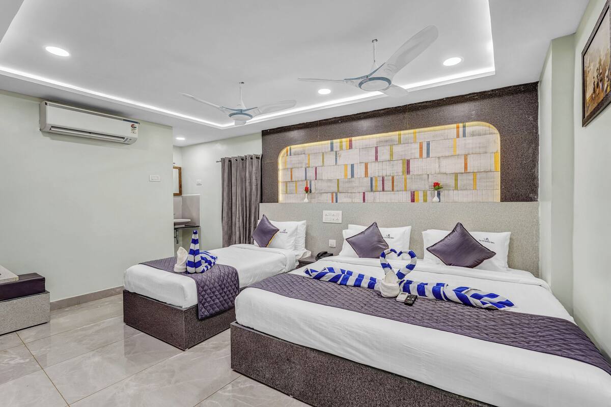 A hotel room with two beds, a ceiling fan, recessed lighting, an air conditioning unit, a decorative wall panel, and a small table with a vase.
