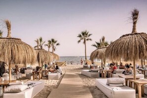 Ein Strand-Loungebereich mit Reetdachschirmen, Liegestühlen und einem Sandweg zum Meer.