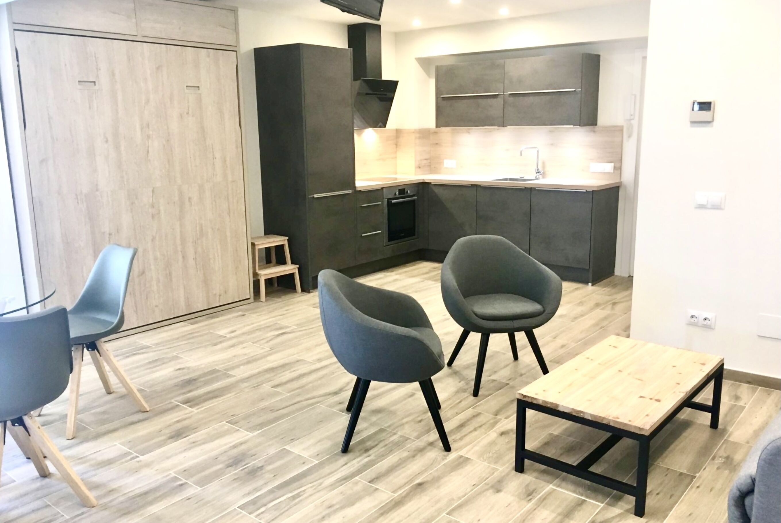 A modern kitchen with dark cabinets, a wooden countertop, and a built-in oven. There are two grey chairs and a small wooden stool.