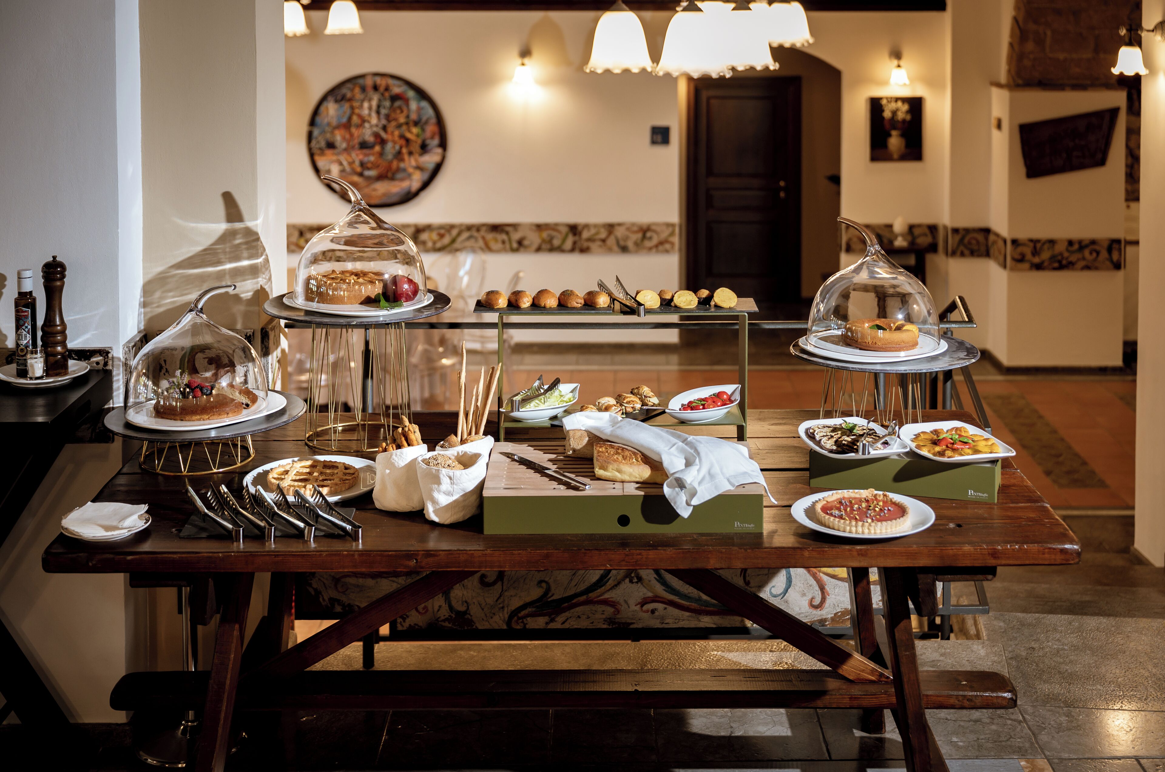 A buffet table with various food items, including pastries and bread, in a well-lit room with a tiled floor and a painting on the wall.