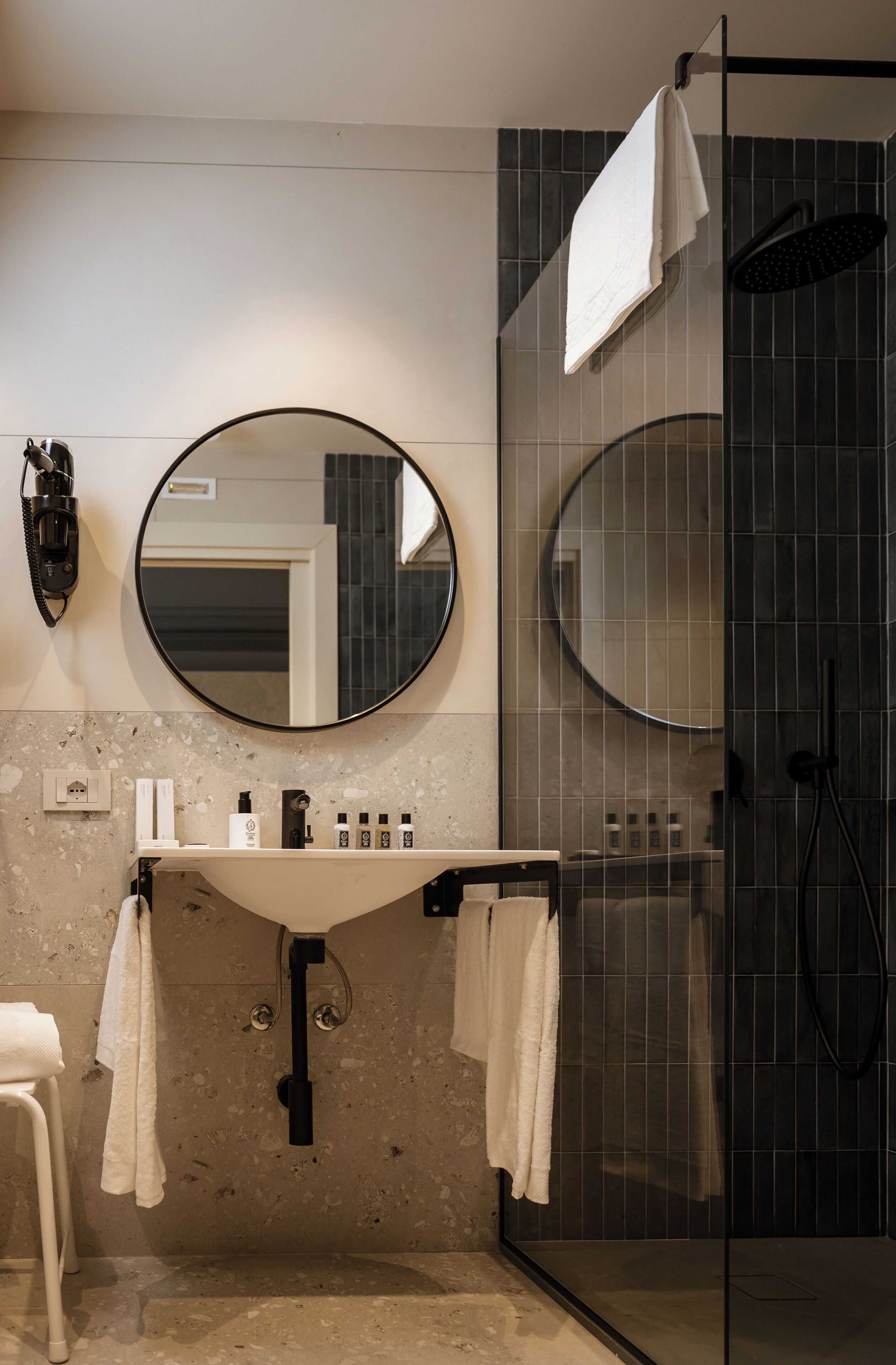 A modern bathroom with a round mirror, a sink with toiletries, and a glass-enclosed shower.