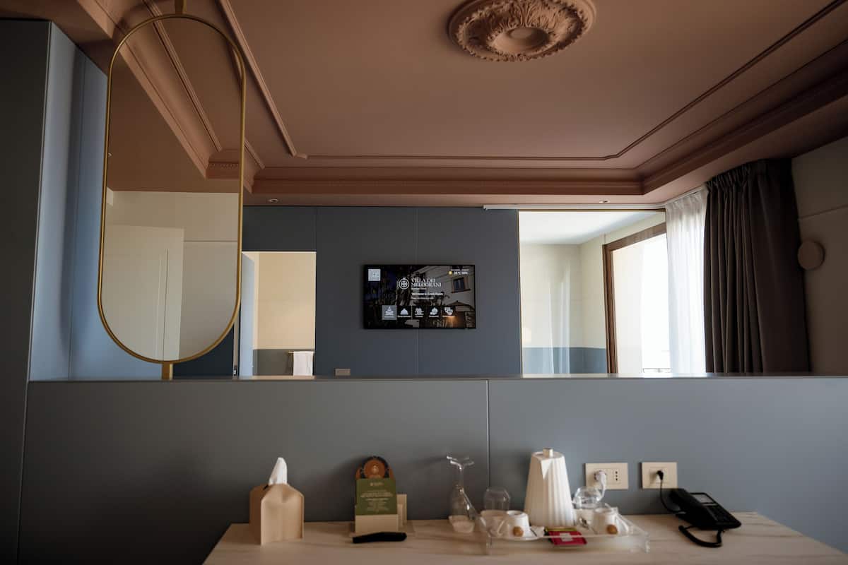 Habitación de hotel con un espejo grande, un televisor de pantalla plana en la pared, una mesita de noche con lámpara, un teléfono y una ventana con cortinas.