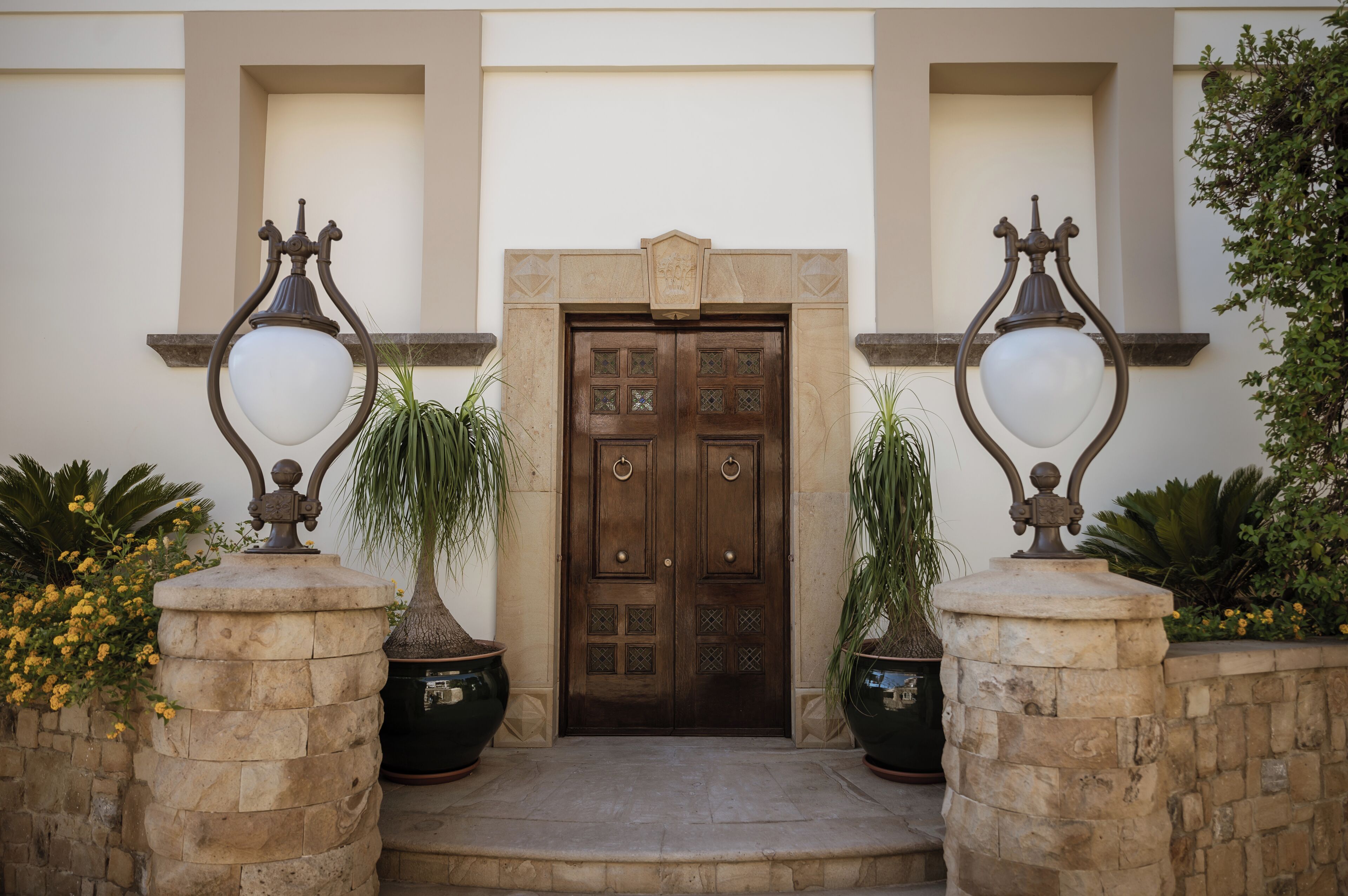 Dos puertas dobles con diseños intrincados, flanqueadas por dos macetas grandes y dos farolas ornamentales.