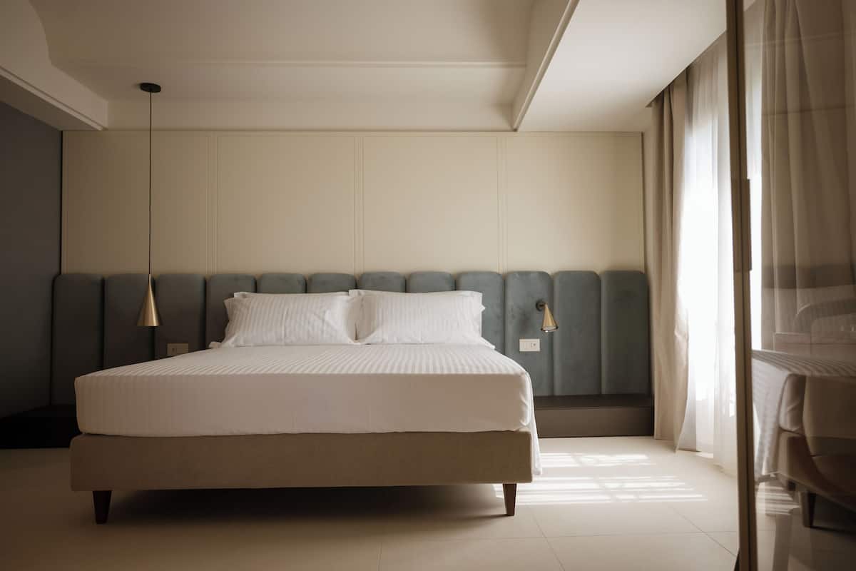 Habitación de hotel moderna con una cama grande, una mesita de noche, una lámpara y vistas al exterior a través de cortinas translúcidas.
