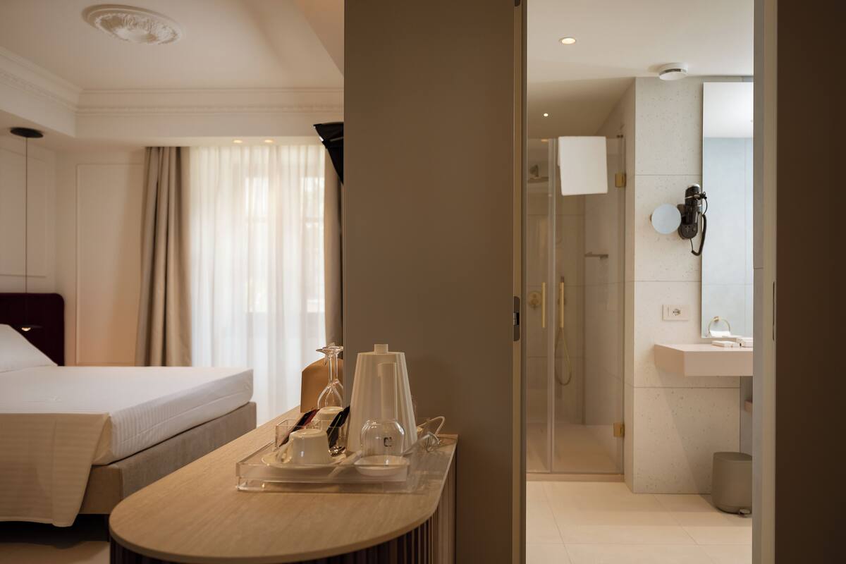Habitación de hotel con cama, mesita de noche con bandeja de desayuno, vaso de agua y zona de ducha con puerta de cristal.