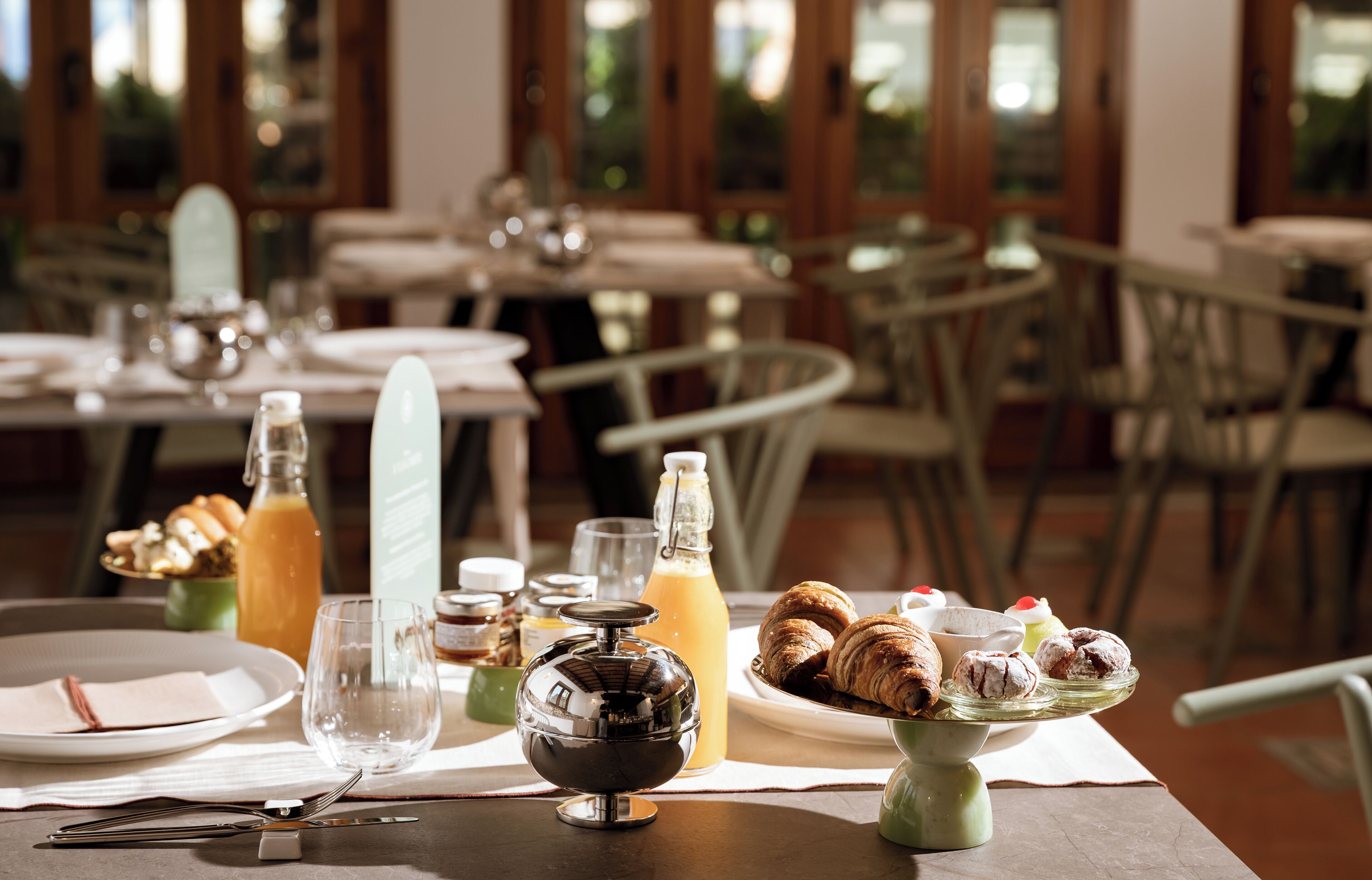 A dining table set with breakfast items, including croissants and pastries, in a restaurant with wooden chairs and a view of greenery through large windows.