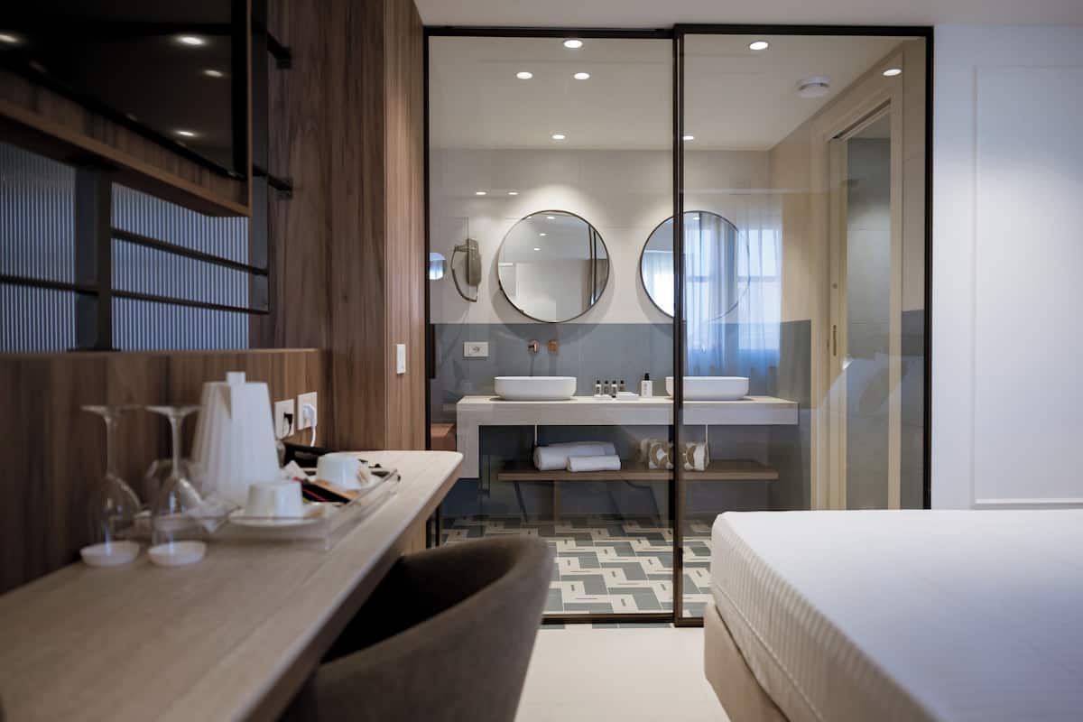 Una habitación de hotel moderna con un escritorio de madera, una cama, un baño con paredes de vidrio que incluye un lavamanos y espejos, y un piso con diseño.