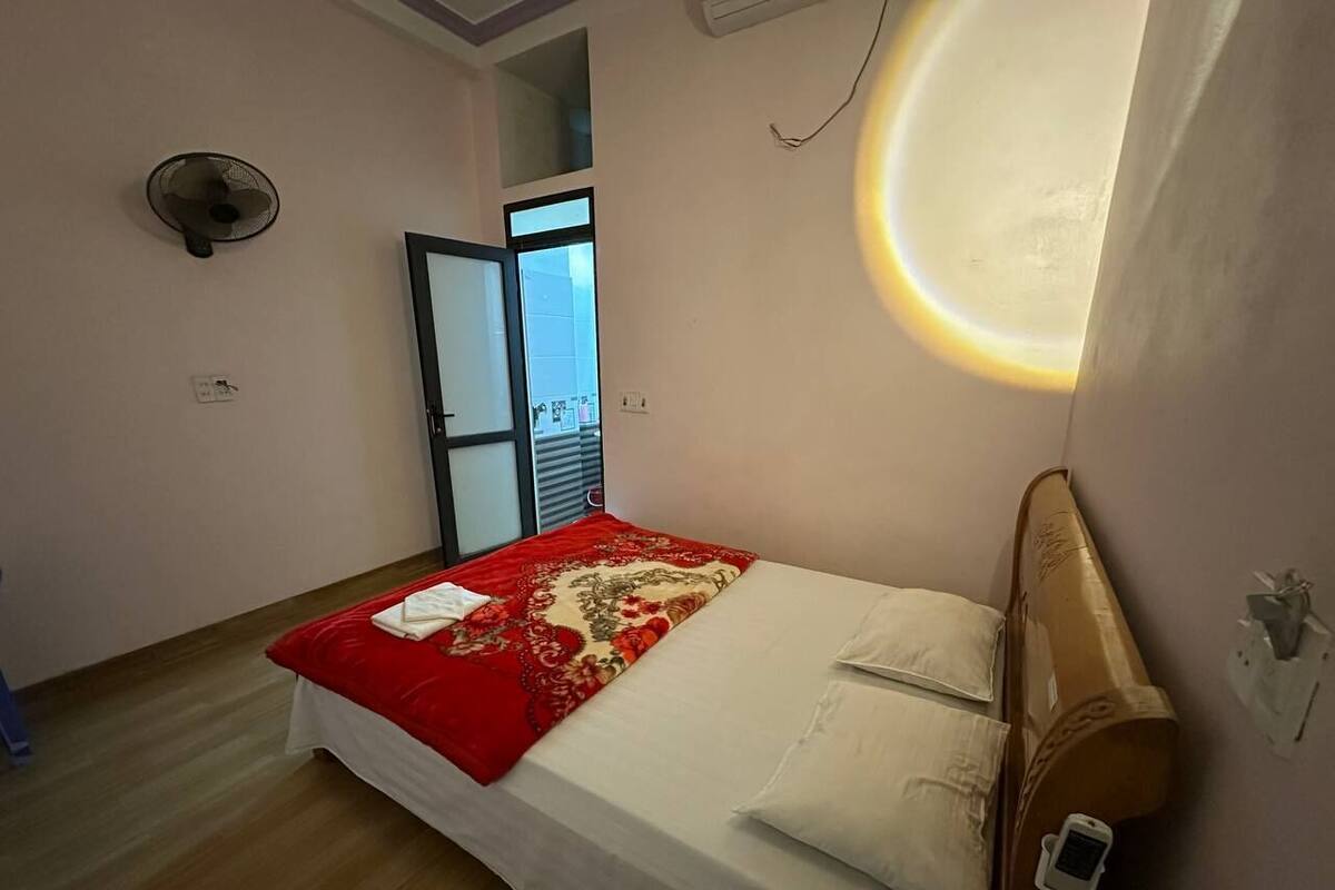 Ein Schlafzimmer mit einem Bett, einer roten und beigen Bettdecke, einem Holzkopfende, einer Wandleuchte und einem Fenster mit Jalousien.