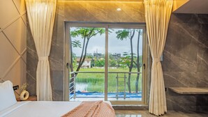 A modern hotel room with a bed, a view of a water body, and a balcony with a railing. - Alaya Stays - Imperial Calista 4BHK near Khandala Hills (Lonavala)