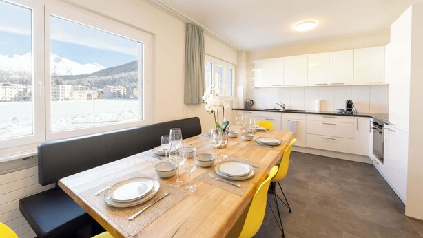 A dining area with a wooden table set for four, a bench, and a view of a snowy mountain landscape through large windows. - Modern Apartment, Near to Skiing Scenic Lake Views (Sankt Moritz)