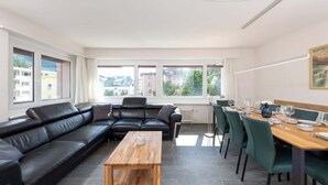 Een moderne woonkamer met een zwarte leren bank, een houten salontafel en een eethoek met groene stoelen en een houten tafel gedekt voor vier personen. Grote ramen bieden uitzicht op de skyline.