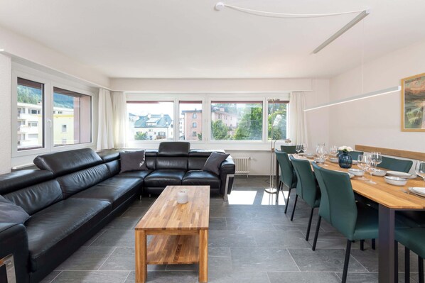 Un salon moderne avec un canapé en cuir noir, une table basse en bois et un coin repas comprenant des chaises vertes et une table en bois, le tout disposé pour accueillir quatre personnes. De grandes fenêtres offrent une vue sur le paysage urbain.