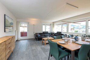 Un salon moderne avec un canapé, une table à manger pour quatre personnes et de grandes fenêtres offrant une vue sur les bâtiments et les montagnes.