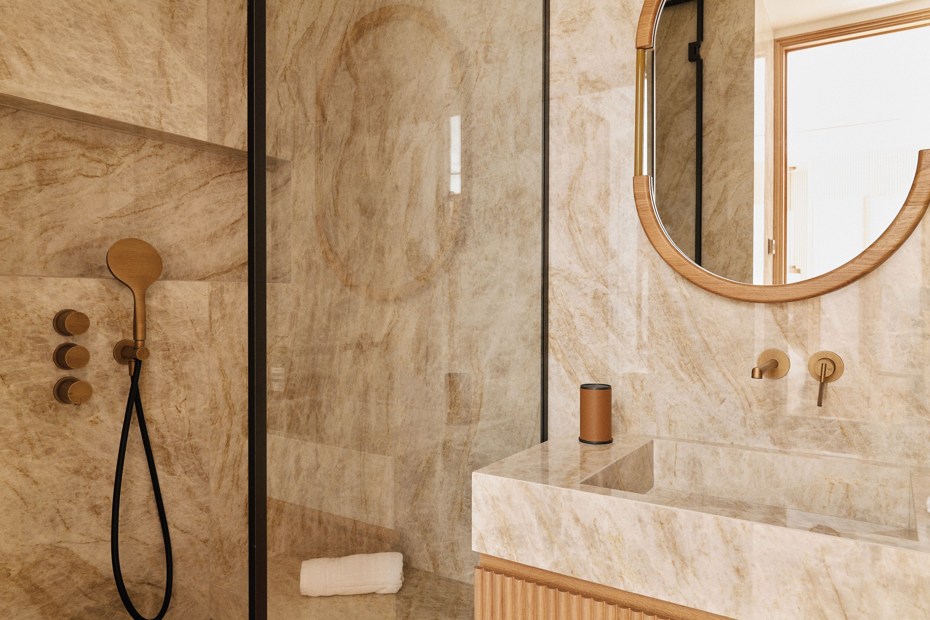 Un baño con pared de mármol, un espejo redondo, una ducha con cabezal de ducha manual y un lavabo con toalla.
